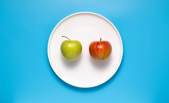 Red and green apple on a white plate on a blue background.