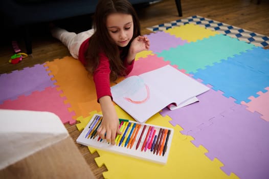 Top view of charming little girl drawing using color pastel crayons while lying on a colorful multicolored puzzle carpet on the floor in her room at home. Kids entertainment and education