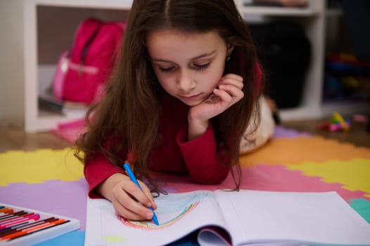 Adorable little child girl drawing rainbow with colorful pastel pencils, lying on a multicolored puzzle carpet in her room at home. Art and creativity concept. Kids development, education. Hobbies and leisure.