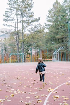 Little girl walks along a sports ground strewn with leaves in an autumn park. Back view. High quality photo