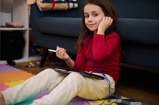 Portrait of a lovely child girl using digital tablet, studying online from home, smiling looking at camera, sitting on colorful puzzle carpet. Kids education. Distance learning. Autism and diversity