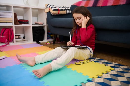 Adorable little child girl putting on earphones, sitting on a multicolored puzzle carpet, with a digital tablet, ready for online lesson. Distance education. Kid entertainment and development concept