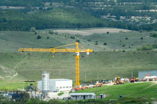 A large crane is on a construction site in a rural area. The crane is yellow and is surrounded by a lot of other construction equipment. The scene is peaceful and quiet