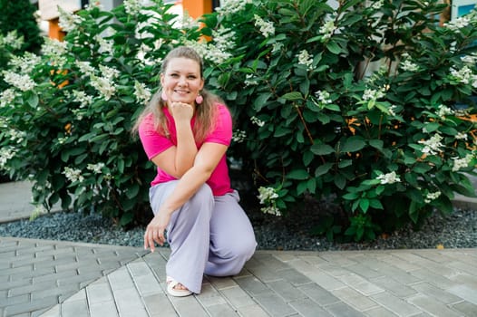 Portrait of beautiful smiling millennial woman wearing stylish pink t shirt looking at camera standing in green park. Positive lifestyle, natural beauty concept. Copy space.