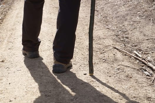 Close-up of a man walking with a wooden cane along a dirt road.