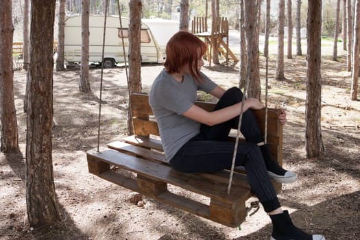 teenage girl sits on a swing made from a wooden pallet in a pine forest.