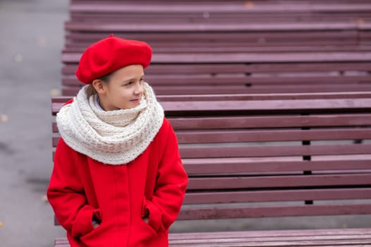 Smiling caucasian girl in a red coat and beret sits alone on a bench
