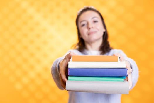 Portrait of smiling woman offering pile of books, recommending reading hobby for relaxation purposes. Radiant bookaholic person with stack of novels in arms doing endorsement, studio background