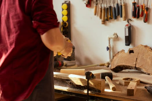 Woodworking specialist using manual sandpaper to sander lumber block, making furniture in assembling shop during job shift. Handyman polishing surface of piece of wood with abrasive material