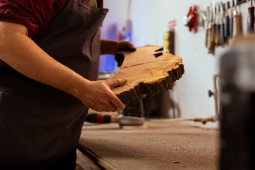 Carpenter holding timber block, doing quality assurance on it before starting furniture assembling in workshop, close up shot. Manufacturer in joinery preparing piece of wood for carving