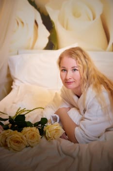 A blonde girl in a white terry bathrobe in hotel room with flowers. Wedding travel, Weekend and vacation, body care