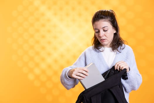 Portrait of woman rummaging through backpack, taking book out, isolated over studio background. Student removing textbook used for educational purposes from school rucksack