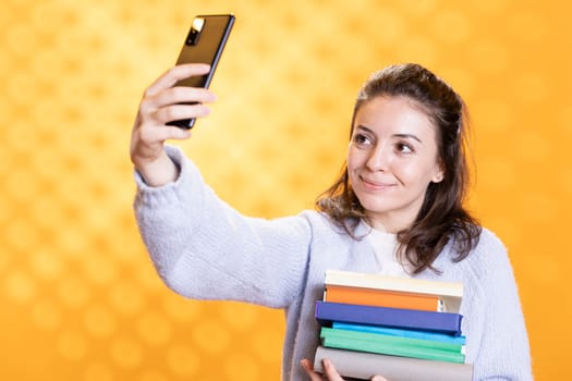 Smiling woman doing selfie with cellphone holding stack of books, isolated over studio background. Happy student with pile of textbooks in arms taking picture with mobile phone