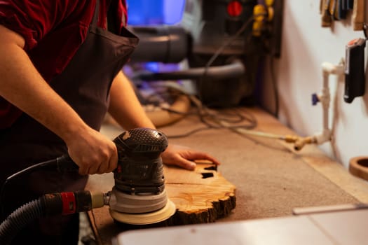 Carpenter holding power tool used to smooth surfaces by abrasion with sandpaper, using it on wood piece. Man using orbital sander equipment for furniture assembling job in studio