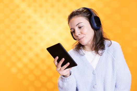 Upbeat woman using ereader to enjoy book, listening music, conveying joy of reading concept, studio background. Geek reading digital novel on tablet screen and hearing songs in headphones