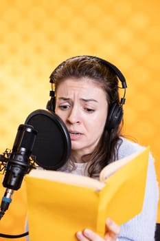 Woman with frown on face wearing headphones reading aloud from book into mic against background. Voice actor recording audiobook, creating engaging content for listeners, glowering for dramatic effect