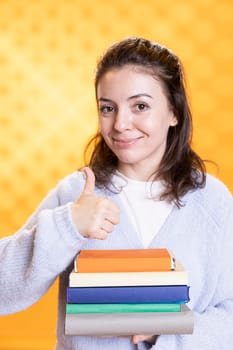 Portrait of cheerful woman carrying stack of books, gathering information for school exam, showing thumbs up, studio background. Happy student holding textbooks, doing positive hand gesture