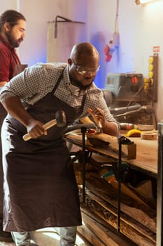 Craftsperson carving designs into wood using chisel and hammer next to coworker. Artisan shaping wooden pieces using protective equipment to prevent workplace accidents, helped by apprentice