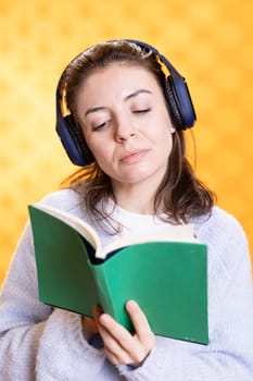 Upbeat woman turning page on book and listening music, conveying joy of reading concept, studio background. Geek reading novel and hearing songs in headphones, showing appreciation for literature