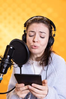 Woman frowns while doing voiceover reading of ebook on ereader to produce audiobook using dramatic acting. Narrator glowers, portraying character, recording novel using digital tablet, studio background