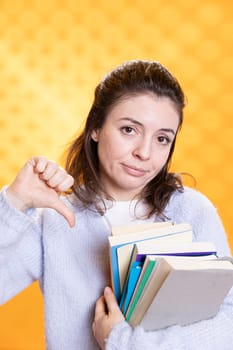 Portrait of woman holding stack of books doing thumbs down sign gesturing, studio background. Stressed student with pile of textbooks in arms used for academic learning doing negative hand gesture