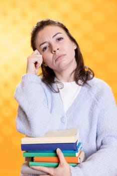 Portrait of pensive woman carrying stack of books, memorizing information for school exam, studio background. Student holding textbooks, remembering and practicing before university assessment