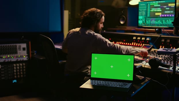 Music production engineer mixing and mastering music in control room, using laptop with greenscreen display. Skilled audio technician working with recording software on pc, pushing faders. Camera B.