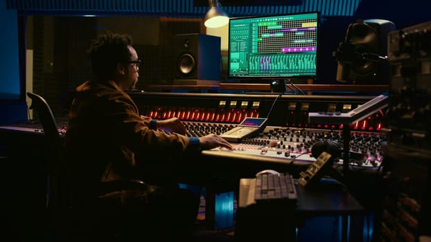 African american sound expert operates on audio console with moving faders and colored meters, editing in professional studio control room. Producer works on album, recording hit song. Camera B.