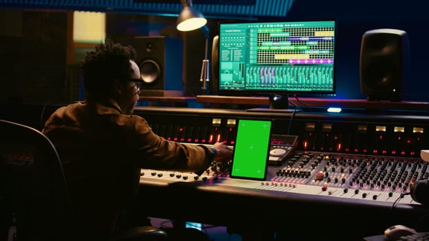 African american audio technician mixing and editing tracks with greenscreen on tablet, working in professional post production studio. Producer creating new tunes for his album. Camera B.