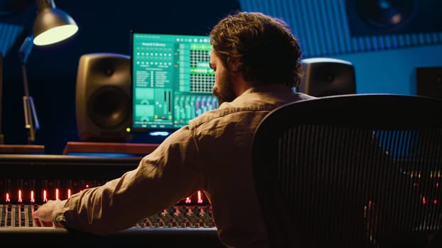 Sound designer mixing and mastering tracks on audio console in control room, using daw software on computer and editing files. Technician creating new music in professional studio. Camera A.