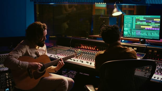 Rockstar recording music on his guitar in professional studio, creating new music for his album in control room. Artist composer producing tracks on electro acoustic guitar. Camera B.