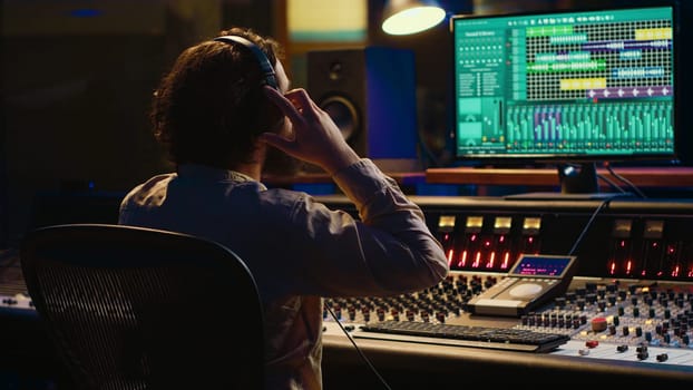Sound designer working on track recording with audio professional software, operating technical equipment on panel board in control room. Audio engineer enhances music high quality. Camera A.