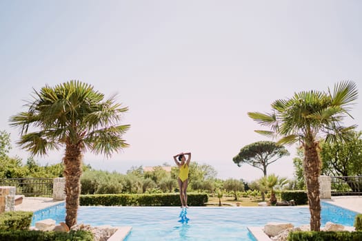 Young woman sunbathing while standing on the edge of the pool, putting her hands behind her head and looking to the side. High quality photo