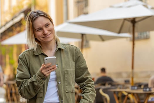 Caucasian young woman using smartphone typing texting social media messages e-mail looking for a way on map in mobile navigator app outdoors. Smiling girl walking on city street. Town lifestyles.