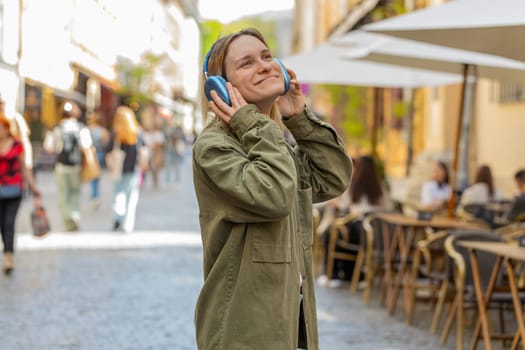 Rear view of young woman tourist with backpack listening to music via headphones walking through street, looking around and smiling. Back view of girl traveler in urban city background. Lifestyles.