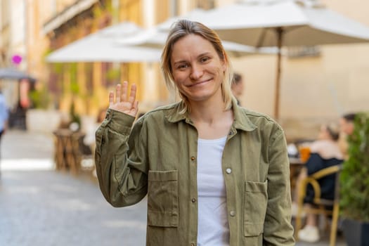 Portrait of girl tourist smiling friendly at camera, waving hands gesturing invitation hello, hi, greeting, goodbye, welcoming with hospitable expression outdoors. Woman on urban city town street.