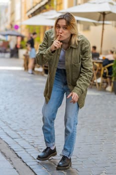 Shh be quiet please. Young woman presses index finger to lips makes silence hush gesture sign do not tells gossip secret outdoors. Caucasian girl tourist standing on urban city street. Town lifestyles