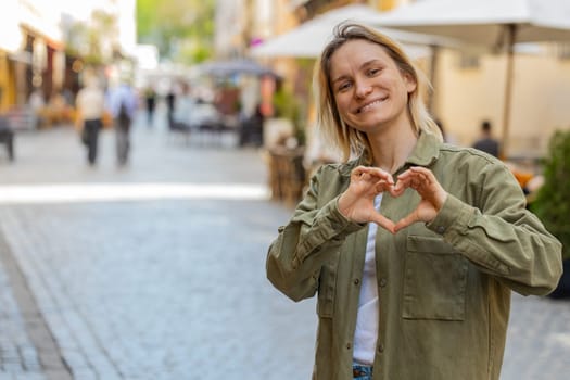 I love you. Young woman makes symbol of love, showing heart sign to camera, express romantic feelings, express sincere positive feelings. Charity, gratitude, donation. Outdoors on urban city street.
