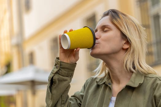 Happy Caucasian young woman enjoying morning coffee hot drink and smiling outdoors. Relaxing, taking a break. Girl walking through urban city center street, drinking coffee to go. Town lifestyles.
