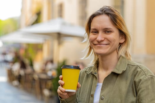 Happy Caucasian young woman enjoying morning coffee hot drink and smiling outdoors. Relaxing, taking a break. Girl walking through urban city center street, drinking coffee to go. Town lifestyles.