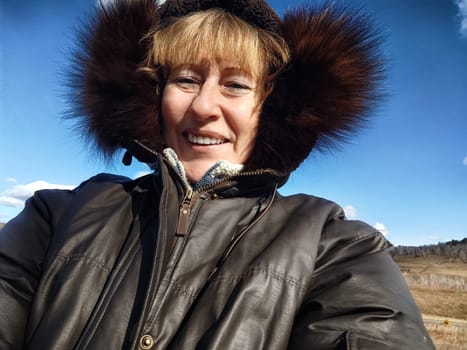 Happy female blonde tourist in a warm jacket taking selfie with outside in cold summer, autumn or winter day. Middle aged woman having fun in nature trip