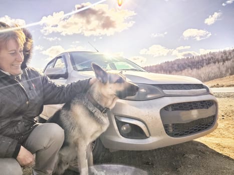 Adult girl with shepherd dog taking selfie near car. Middle aged woman and big pet in travel on nature. Friendship, love, fun and hugs