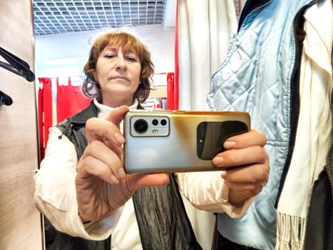 A middle-aged blonde woman in a cramped fitting room of a store with a bunch of clothes and hangers takes selfie in the mirror