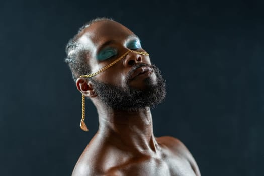 Portrait of shirtless African American gay man with blue eyeshadows and golden chain on face with closed eyes against dark blue background