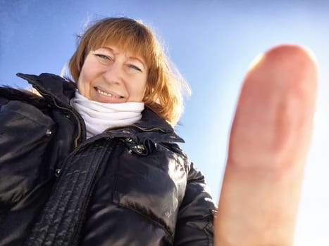 A cheerful middle aged woman in a winter coat taking selfie on nature outdoors in sunny day with blue sky