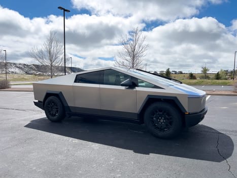 Denver, Colorado, USA-March 28, 2024-Captured from the side, a Tesla Cybertruck stands prominently in a vast parking lot, reflecting the innovation of electric vehicles against a serene, natural backdrop.