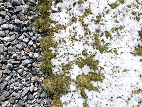 An image showing the remnants of a spring snowstorm on a suburban landscape, where melting snow meets the contrasting textures of gravel and green grass.