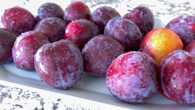A selection of juicy, ripe plums are artfully arranged on a plate, resting on a marble countertop in a bright, modern kitchen, awaiting to be enjoyed.