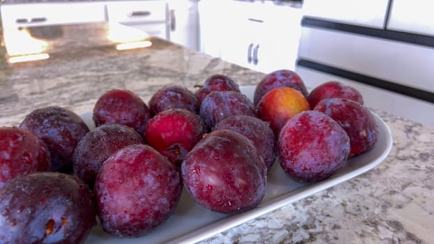 A selection of juicy, ripe plums are artfully arranged on a plate, resting on a marble countertop in a bright, modern kitchen, awaiting to be enjoyed.