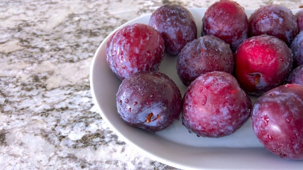 A selection of juicy, ripe plums are artfully arranged on a plate, resting on a marble countertop in a bright, modern kitchen, awaiting to be enjoyed.